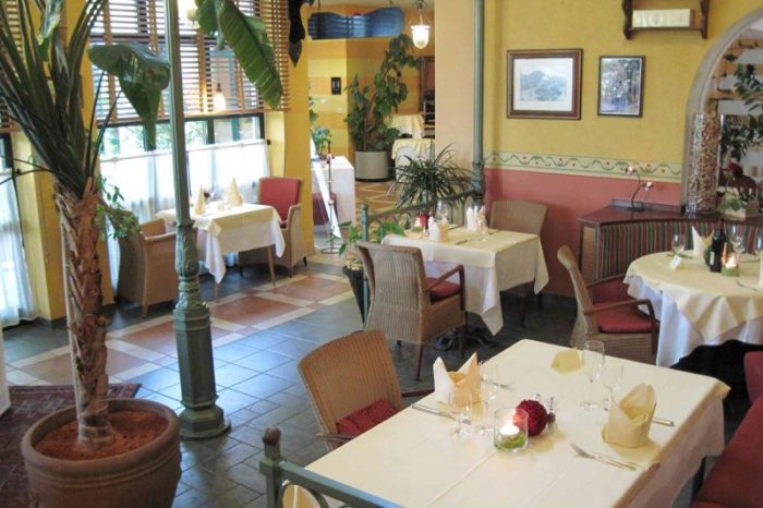 Lichterfüllter Restaurant-Wintergarten mit hohen Decken, Pflanzen und mediterranem Ambiente.