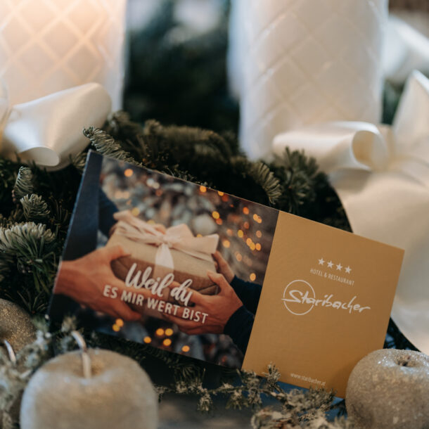 Ein Gutschein vom Hotel Staribacher vor einem Adventkranz mit weißen Kerzen.