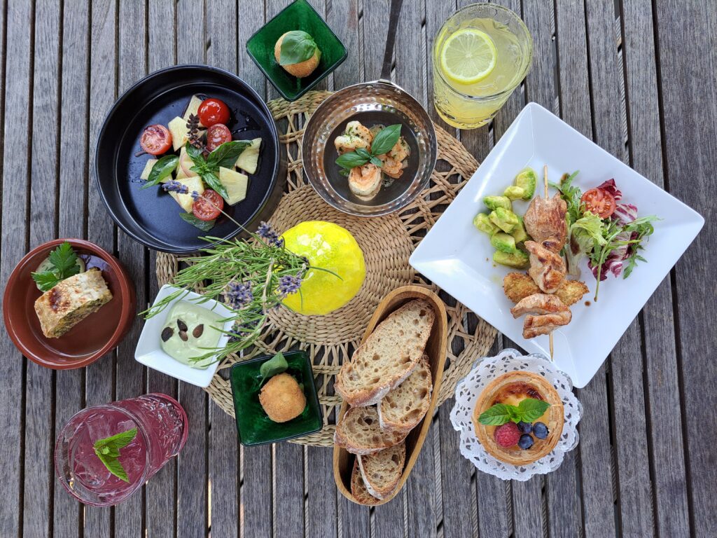 Eine appetitliche Draufsicht (Flat Lay) auf einen rustikalen Holztisch, der reich gedeckt ist mit verschiedenen Tellern und Platten einer sommerlichen Bistro-Tafel. Im Zentrum steht eine Holzplanke mit Stücken eines knusprigen Flammkuchens, reich belegt mit karamellisierten roten Zwiebeln, Lauch, gebratenem Speck und gerösteten Kürbiskernen. Drumherum sind ein schwarzer Teller mit frischem Caprese-Salat (Kirschtomaten, Mozzarella, Basilikum, rote Zwiebeln), ein weißer Teller mit einem gegrillten Hähnchenspieß auf Avocadowürfeln und Tomaten sowie ein kleiner, süßer Tartelette mit Beeren-Topping (Himbeere, Blaubeere) auf einem Spitzendeckchen angeordnet. Ein Glas mit eiskalter, violetter Limonade und frischer Minze sowie Brotstücke sind ebenfalls zu sehen. Die Farben sind lebendig und appetitlich.