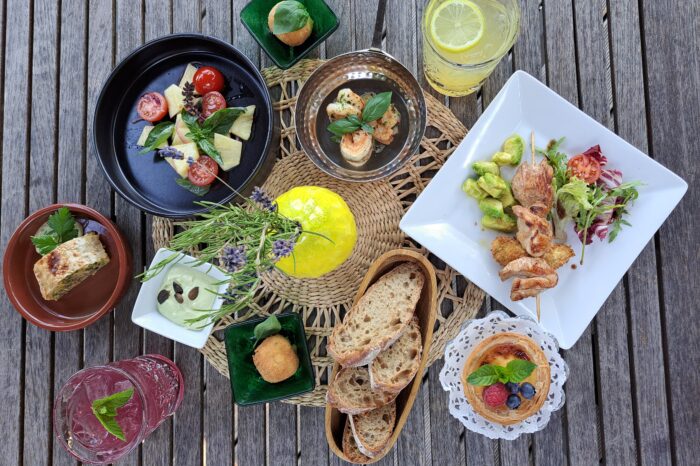 Eine appetitliche Draufsicht (Flat Lay) auf einen rustikalen Holztisch, der reich gedeckt ist mit verschiedenen Tellern und Platten einer sommerlichen Bistro-Tafel. Im Zentrum steht eine Holzplanke mit Stücken eines knusprigen Flammkuchens, reich belegt mit karamellisierten roten Zwiebeln, Lauch, gebratenem Speck und gerösteten Kürbiskernen. Drumherum sind ein schwarzer Teller mit frischem Caprese-Salat (Kirschtomaten, Mozzarella, Basilikum, rote Zwiebeln), ein weißer Teller mit einem gegrillten Hähnchenspieß auf Avocadowürfeln und Tomaten sowie ein kleiner, süßer Tartelette mit Beeren-Topping (Himbeere, Blaubeere) auf einem Spitzendeckchen angeordnet. Ein Glas mit eiskalter, violetter Limonade und frischer Minze sowie Brotstücke sind ebenfalls zu sehen. Die Farben sind lebendig und appetitlich.