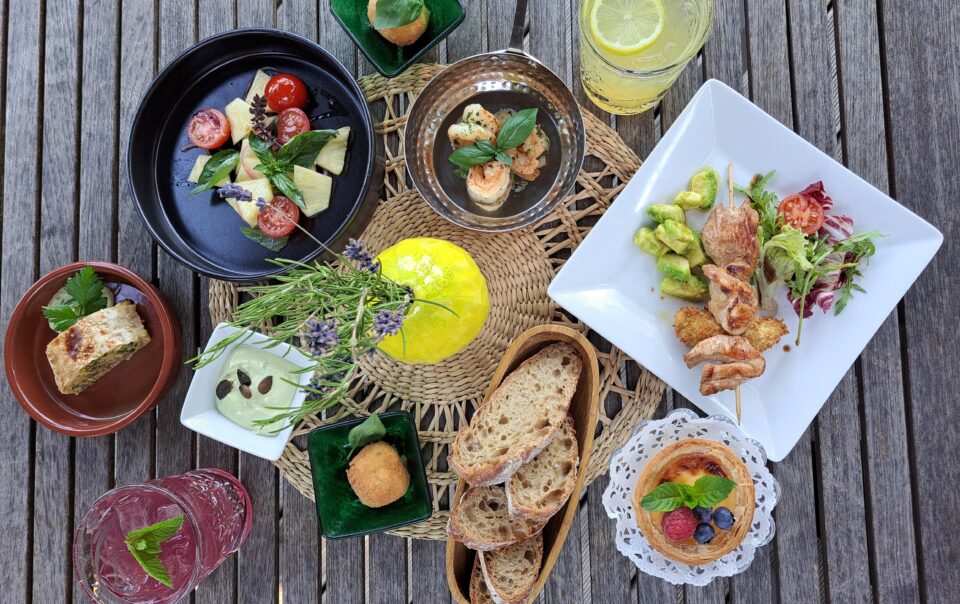 Eine appetitliche Draufsicht (Flat Lay) auf einen rustikalen Holztisch, der reich gedeckt ist mit verschiedenen Tellern und Platten einer sommerlichen Bistro-Tafel. Im Zentrum steht eine Holzplanke mit Stücken eines knusprigen Flammkuchens, reich belegt mit karamellisierten roten Zwiebeln, Lauch, gebratenem Speck und gerösteten Kürbiskernen. Drumherum sind ein schwarzer Teller mit frischem Caprese-Salat (Kirschtomaten, Mozzarella, Basilikum, rote Zwiebeln), ein weißer Teller mit einem gegrillten Hähnchenspieß auf Avocadowürfeln und Tomaten sowie ein kleiner, süßer Tartelette mit Beeren-Topping (Himbeere, Blaubeere) auf einem Spitzendeckchen angeordnet. Ein Glas mit eiskalter, violetter Limonade und frischer Minze sowie Brotstücke sind ebenfalls zu sehen. Die Farben sind lebendig und appetitlich.
