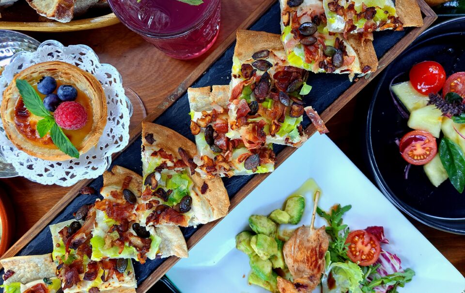 Eine appetitliche Draufsicht (Flat Lay) auf einen rustikalen Holztisch, der reich gedeckt ist mit verschiedenen Tellern und Platten einer sommerlichen Bistro-Tafel. Im Zentrum steht eine Holzplanke mit Stücken eines knusprigen Flammkuchens, reich belegt mit karamellisierten roten Zwiebeln, Lauch, gebratenem Speck und gerösteten Kürbiskernen. Drumherum sind ein schwarzer Teller mit frischem Caprese-Salat (Kirschtomaten, Mozzarella, Basilikum, rote Zwiebeln), ein weißer Teller mit einem gegrillten Hähnchenspieß auf Avocadowürfeln und Tomaten sowie ein kleiner, süßer Tartelette mit Beeren-Topping (Himbeere, Blaubeere) auf einem Spitzendeckchen angeordnet. Ein Glas mit eiskalter, violetter Limonade und frischer Minze sowie Brotstücke sind ebenfalls zu sehen. Die Farben sind lebendig und appetitlich.